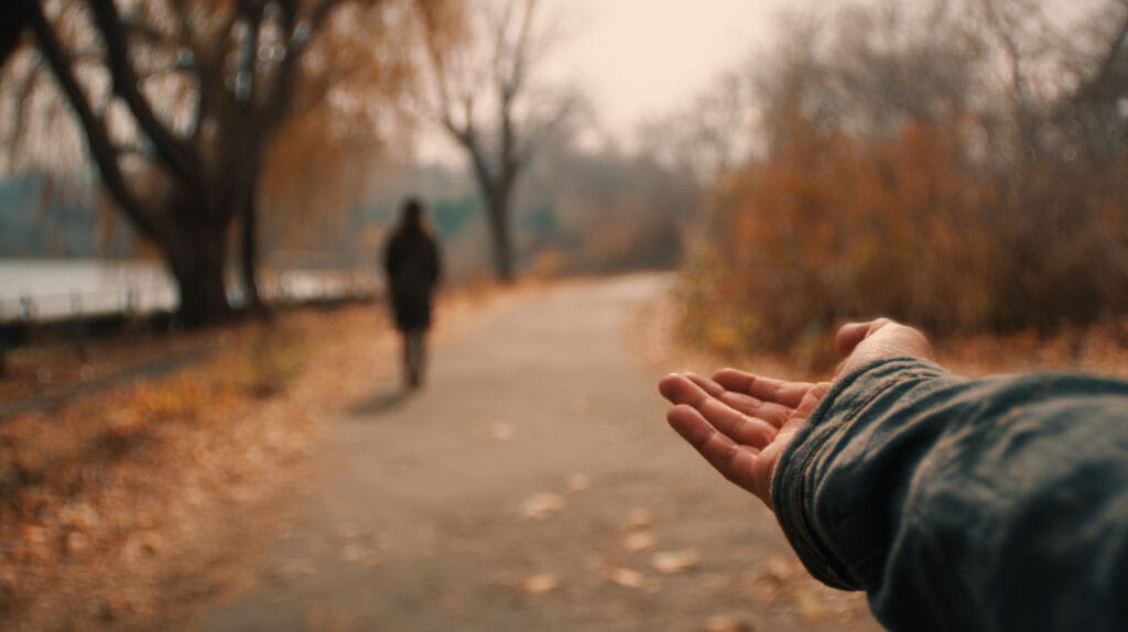 A concerned family member waiting beside a park path with an open hand extended toward a loved one approaching from a distance